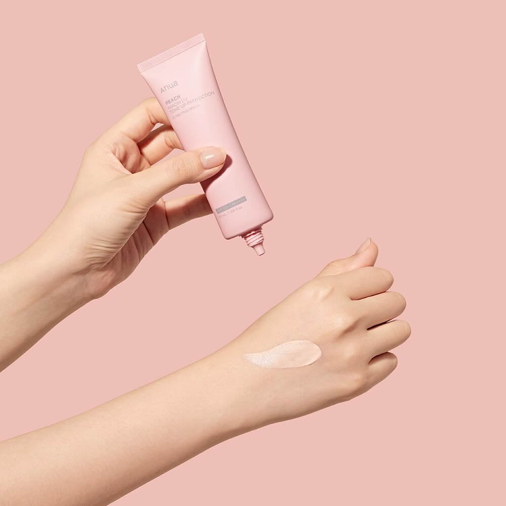 Anua Peach Niacin UV Tone Up Protection 01 Melting Peach applied to hand with pink background, showcasing hydrating sunscreen.