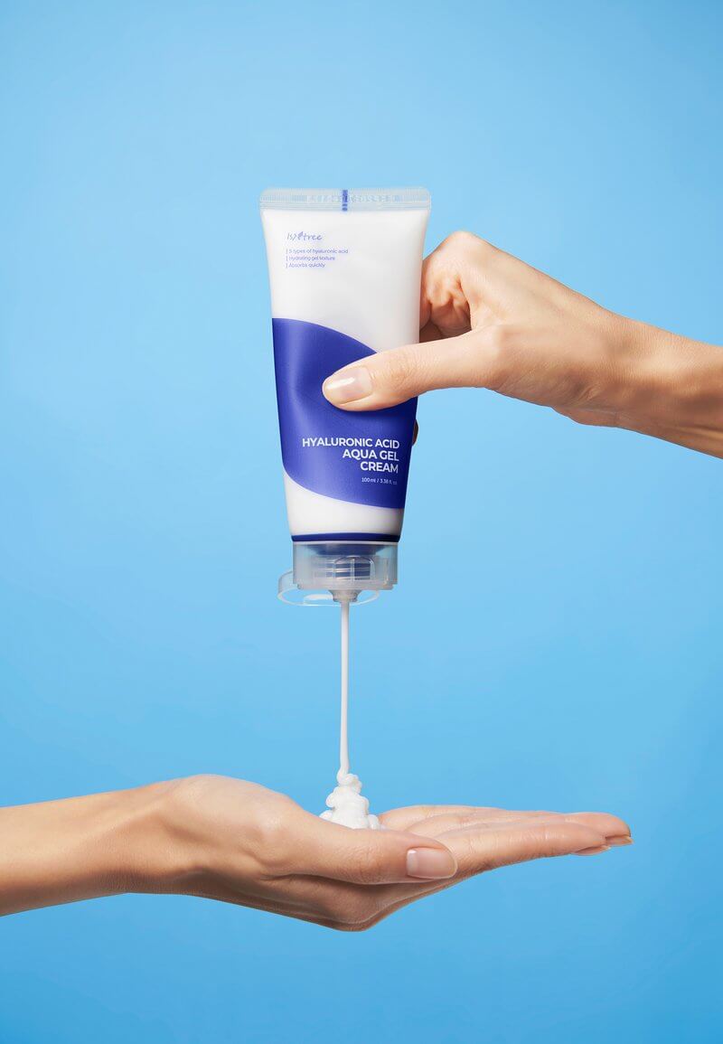 Hand holding Isntree Hyaluronic Acid Aqua Gel Cream tube, dispensing moisturizing gel on palm against blue background.