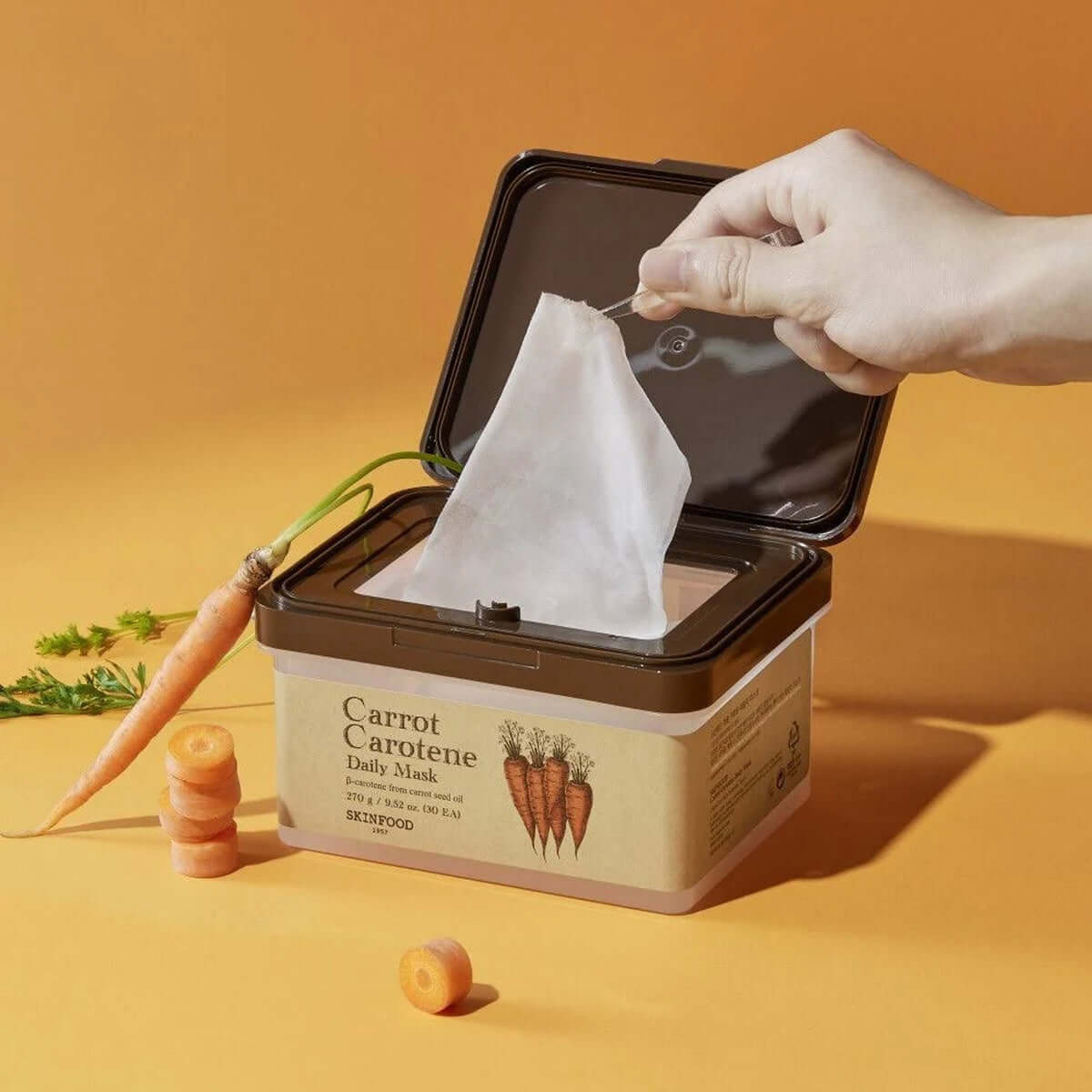 SKINFOOD Carrot Carotene Daily Mask packaging with a hand holding a mask sheet next to carrots.