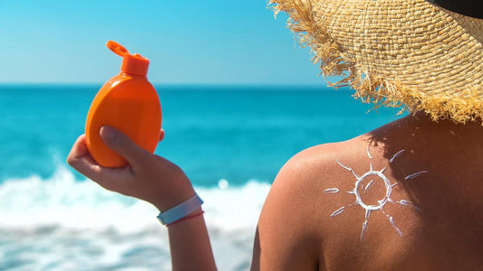 Person holding sunscreen bottle with sun drawn on shoulder at the beach, emphasizing importance of SPF for skin protection.
