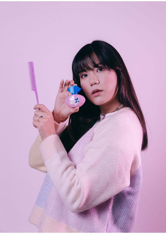 Woman in pastel sweater holding a cute hair product and a comb against a pink background.