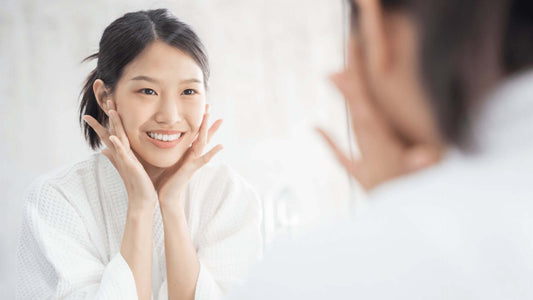 Woman in bathrobe smiling and applying skincare while looking in the mirror, highlighting Korean skin health.